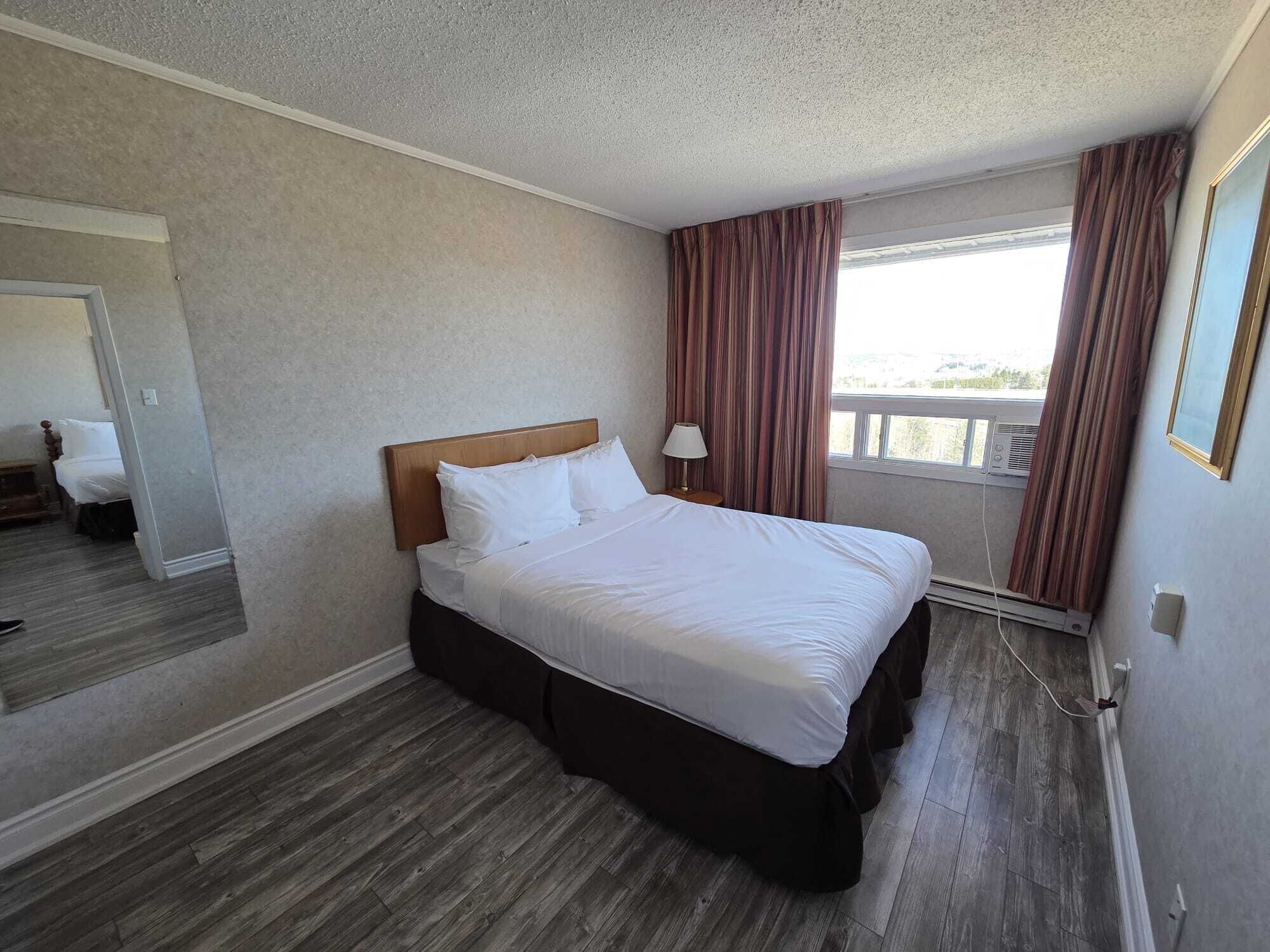 A hotel room with a bed, a window with curtains, a bedside table with a lamp, and a view of the outside.