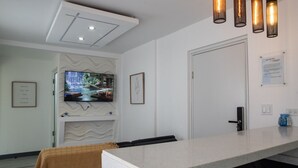 Habitación de hotel moderna con televisión de pantalla plana, techo blanco con iluminación empotrada, una puerta y una pequeña mesa con un objeto azul encima.