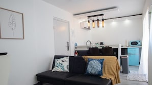 Una sala moderna con un sofá negro, un tapete estampado, una zona de cocina con un refrigerador azul y una lámpara de techo.