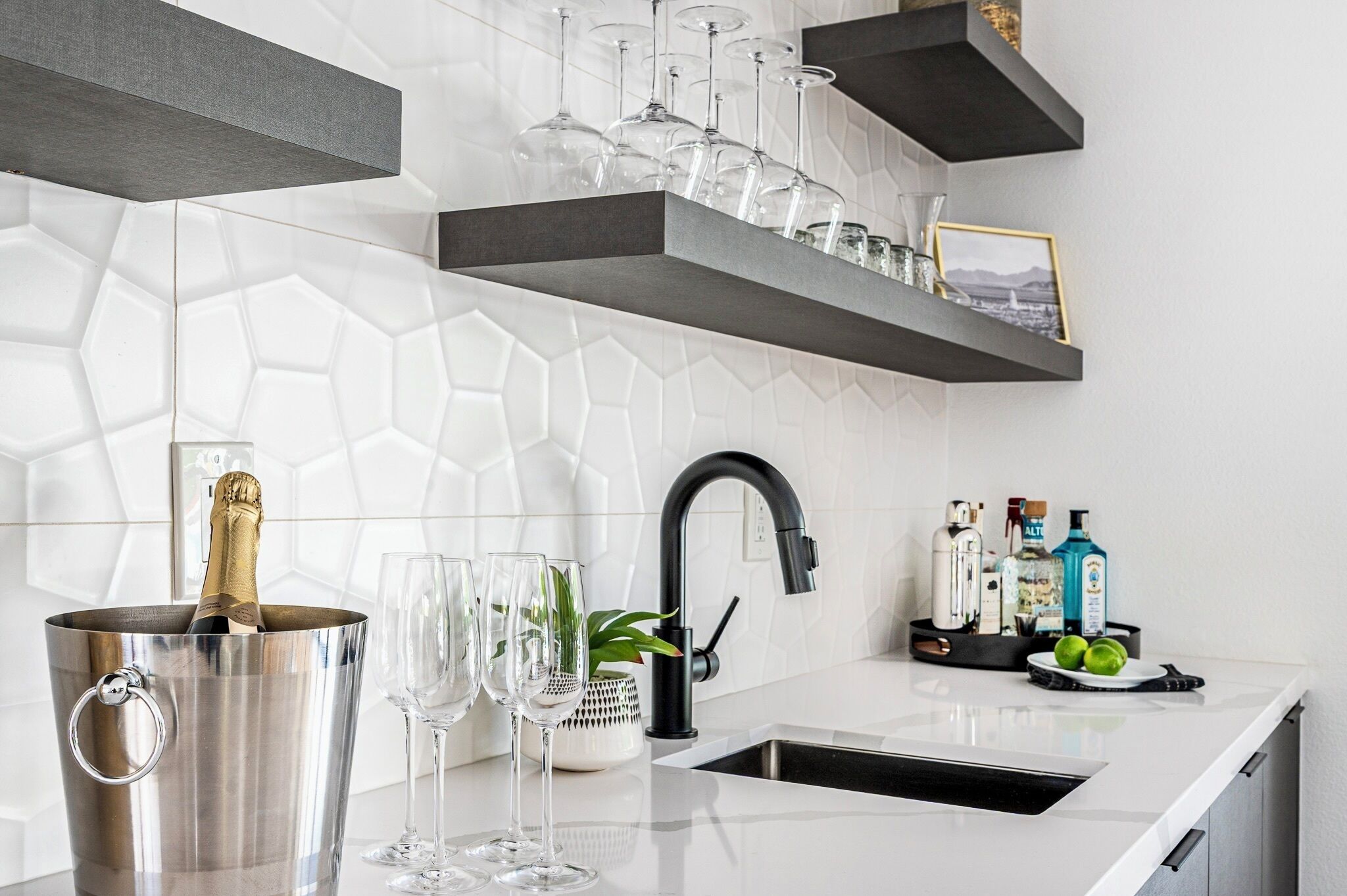 Une cuisine moderne avec un plan de travail blanc, un robinet noir et un porte-bouteilles contenant des verres. On aperçoit une bouteille de champagne dans un seau en métal et une petite plante sur le plan de travail.