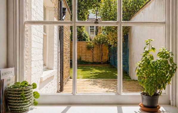 Une vue à travers une fenêtre donnant sur une terrasse en bois, un mur de briques et un jardin.