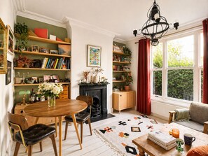 Un salon chaleureux avec une cheminée, des étagères à livres, un coin repas et une grande fenêtre avec des rideaux.