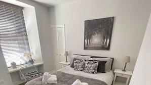 A bedroom with a bed, two bedside tables, a window with blinds, a wall-mounted artwork, and a radiator. - High Street House Sleeps 4 with Parking (South Lanarkshire)