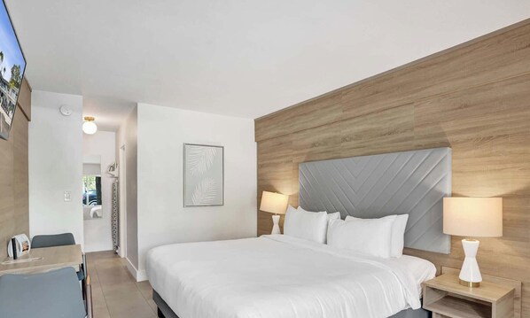 Quarto de hotel moderno com uma cama grande, cabeceira de madeira, duas luminárias laterais, uma obra de arte emoldurada na parede e uma pequena mesa com abajur.