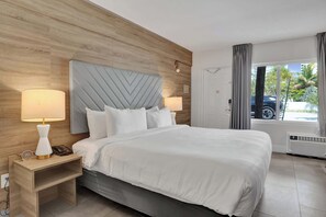 A hotel room with a large bed, two bedside lamps, a wooden headboard, a nightstand, and a view of the outdoors. - Ocean Blvd's Bright King Suite (Fort Lauderdale)