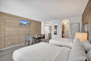 Ein modernes Hotelzimmer mit einer Wandpaneele aus Holz, einem Flachbildfernseher, einem Schreibtisch mit Stühlen, einem Bett mit Kissen, einem Nachttisch mit Lampe und einem Spiegel.