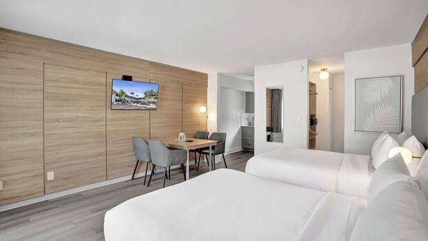 A modern hotel room with a wooden accent wall, a dining area with a table and chairs, a bed with white bedding, and a TV mounted on the wall. - Ocean Blvd Double Suite with Kitchenette at The Botanica! (Fort Lauderdale)