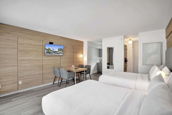 Ein modernes Hotelzimmer mit einer Wand in Holzoptik, einer Essecke mit Tisch und Stühlen, einem Bett mit weißer Bettwäsche und einem an der Wand befestigten Fernseher.