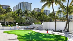 A golf putting green with white and red flags, surrounded by palm trees and modern buildings. - Ocean Blvd Double Suite with Kitchenette at The Botanica! (Fort Lauderdale)