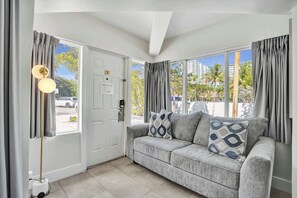 Ein Zimmer mit grauem Sofa, stehender Stehlampe und einer Tür, durch die man Palmen und Gebäude sieht.