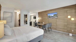 A modern hotel room with a bed, a dining table with chairs, a TV mounted on the wall, and a kitchen area in the background.