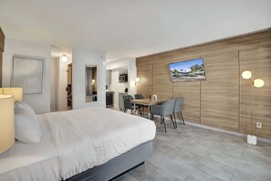 Ein modernes Hotelzimmer mit einem Bett, einem Esstisch mit Stühlen, einem an der Wand montierten Fernseher und einer Küchenzeile im Hintergrund.