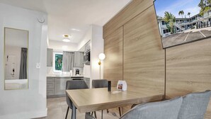 A modern hotel room with a wooden accent wall, a dining area with a table and chairs, a kitchen in the background, and a view of the hotel exterior.