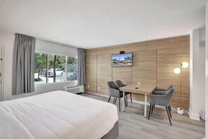 Ein Hotelzimmer mit Bett, Schreibtisch mit zwei Stühlen, Wandfernseher und großem Fenster mit Vorhängen.