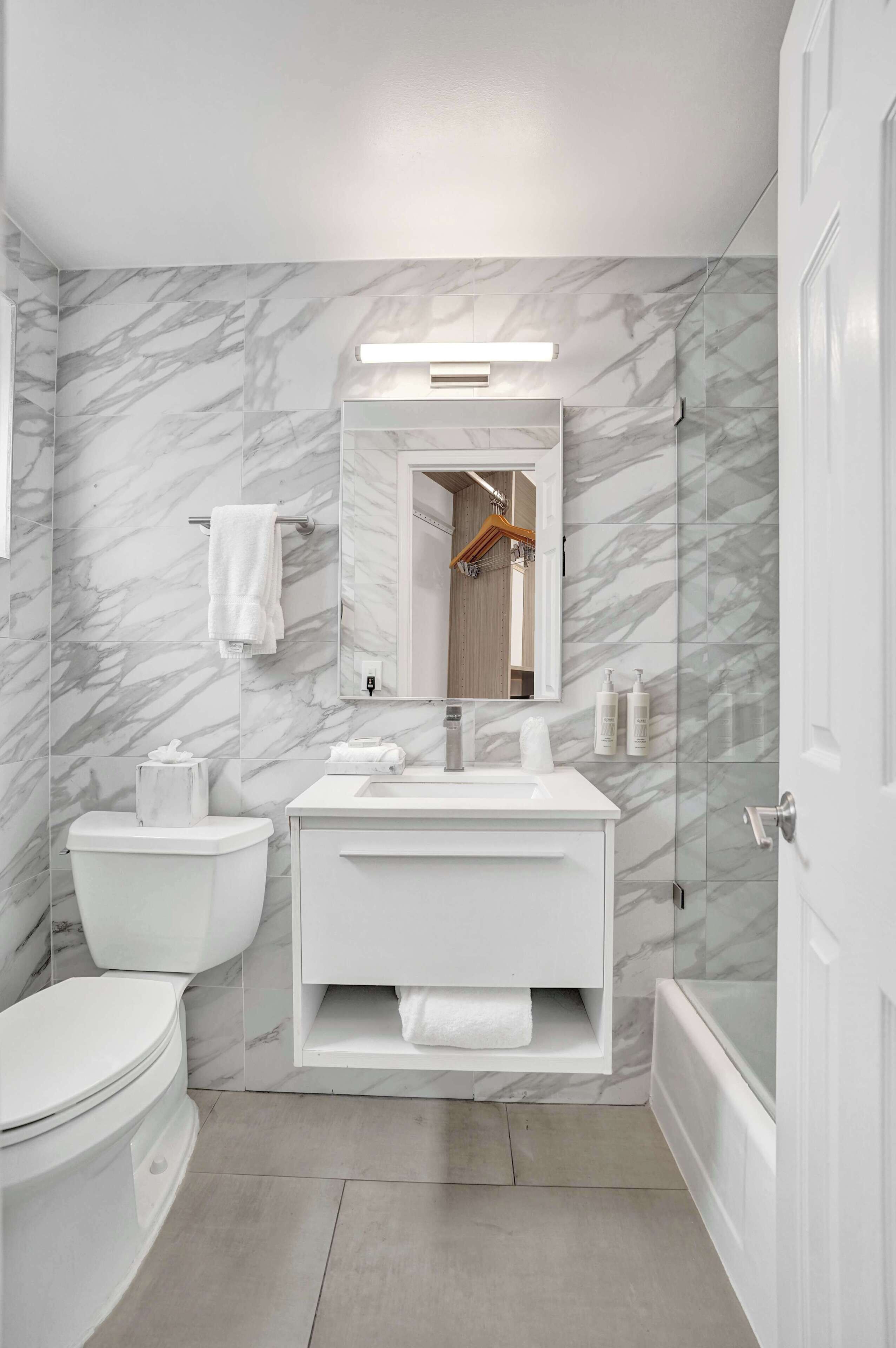 Een moderne badkamer met een marmeren wand, een wit wastafelmeubel, een toilet en een douchecabine van glas.