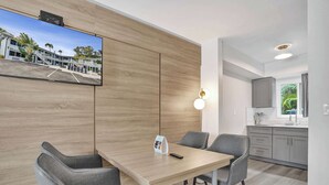 A modern hotel room with a wooden accent wall, a flat-screen TV, a dining area with grey chairs, and a kitchen with grey cabinets. - Boutique King Suite w/ Kitchenette (Fort Lauderdale)