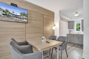 A modern hotel room with a wooden accent wall, a flat-screen TV, a dining area with grey chairs, and a kitchen with grey cabinets. - Boutique King Suite w/ Kitchenette (Fort Lauderdale)