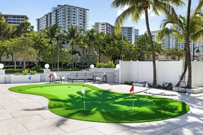 Un campo de golf con un green de putting, palmeras y edificios modernos al fondo.