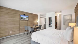 A modern hotel room with a large bed, a desk with chairs, a TV mounted on the wall, and a view of the outside. - Boutique King Suite w/ Kitchenette (Fort Lauderdale)