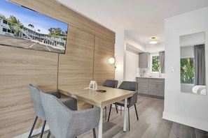 Ein modernes Hotelzimmer mit Holzwand, Flachbildfernseher, Esstisch mit Stühlen, Küchenzeile und großem Fenster.