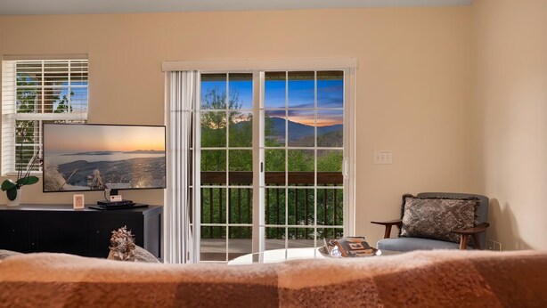 Una habitación con una televisión sobre un pedestal negro, una ventana con persianas, una silla con cojín y vistas a un paisaje desde la ventana.