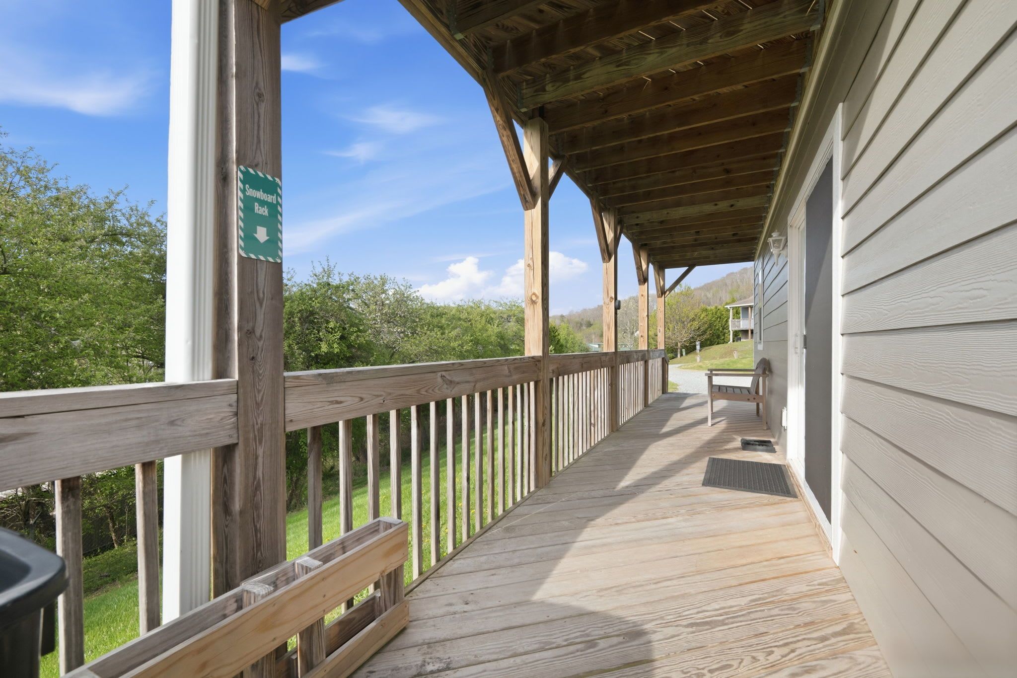 A wooden deck with a sign indicating a snowboard rack, a bench, and a view of greenery.