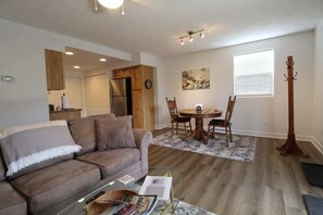 Ein Wohnzimmer mit Sofa, einem Esstisch mit Stühlen, einem Garderobenständer und einer Küchenzeile mit Holzschränken und einem Kühlschrank.