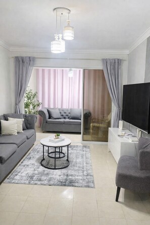 A modern living room with a grey sofa, a round coffee table, a TV, and a glass door leading to a balcony. - Cozy Apt | WiFi +AC | Great Location| Easy Checkin (Santo Domingo)