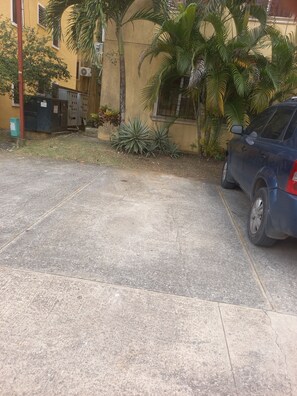 A parking area with a concrete surface, a parked car, and a building with a yellow facade and palm trees. - Cozy Apt | WiFi +AC | Great Location| Easy Checkin (Santo Domingo)