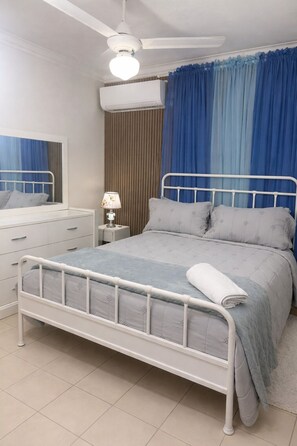 A bedroom with a bed, bedside table, lamp, air conditioning unit, curtains, and a ceiling fan. - Cozy Apt | WiFi +AC | Great Location| Easy Checkin (Santo Domingo)