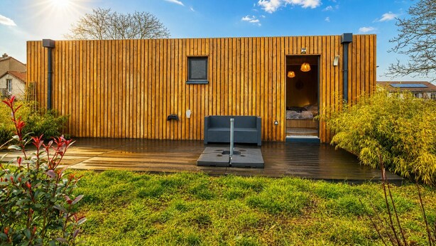 Een moderne houten blokhut met een klein terras, een bank en een bed dat van binnen zichtbaar is.