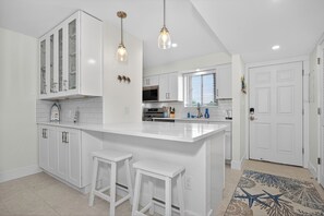 A modern kitchen with white cabinetry, a central island, bar stools, pendant lighting, and a patterned rug. - 
NEW Gulf‑Front 2BR-2BA - Direct View of Pool-Beach-Gulf (Fort Myers Beach)