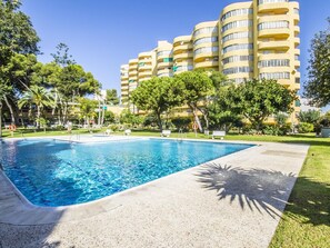 A swimming pool with a clear blue water surface, surrounded by a paved area with benches. In the background, there are multi-story buildings with balconies and a well-maintained lawn with trees. - El Rocio 3B1 - 2 Bedrooms, very close to the beach, swimming pool (Torremolinos)