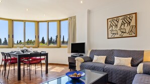 En moderne stue med sofa, sofabord i glas, spiseområde med stole, fjernsyn og store vinduer med udsigt.