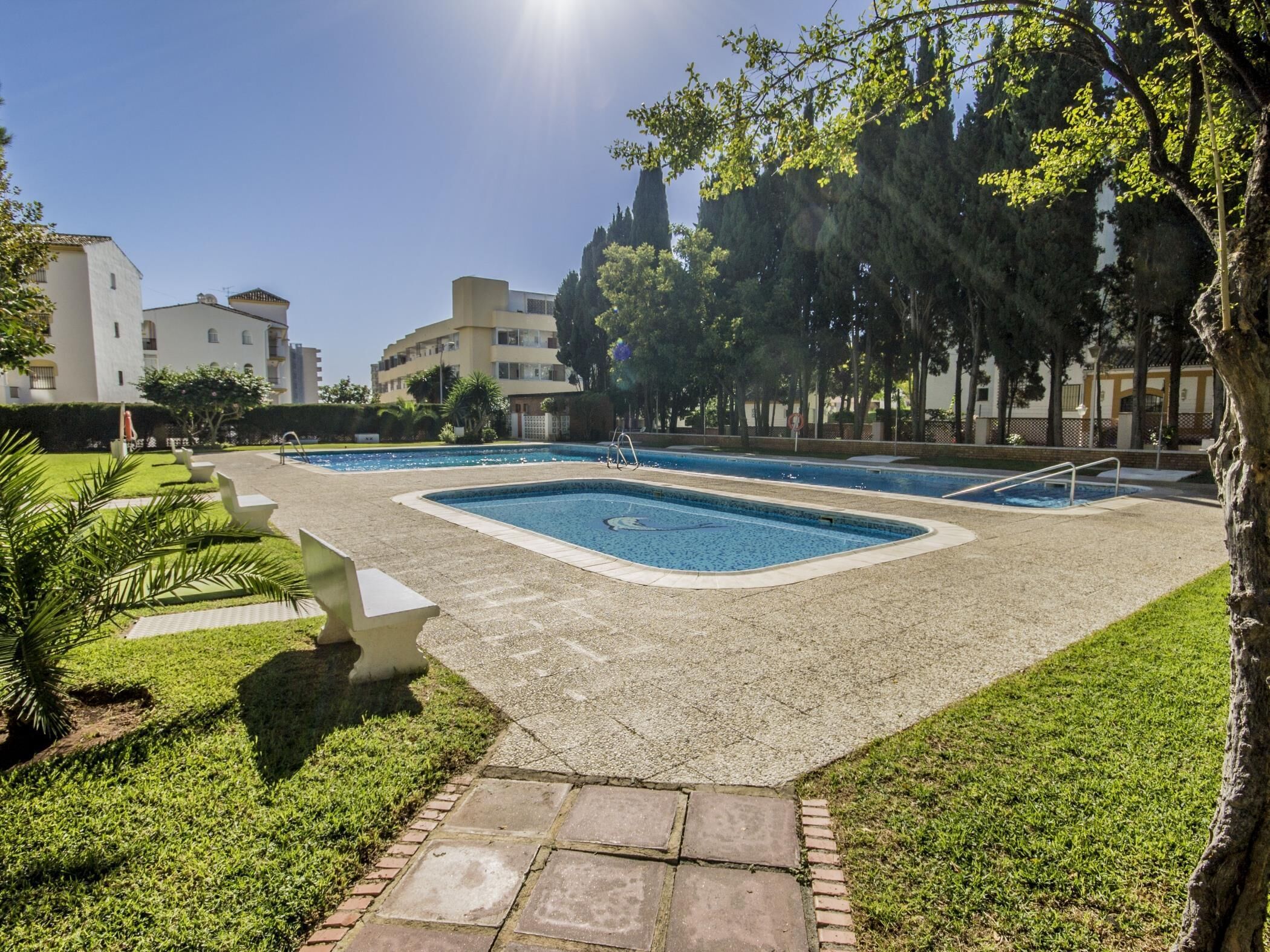 A swimming pool area with two pools, surrounded by a paved walkway, and adjacent to a building with multiple floors.