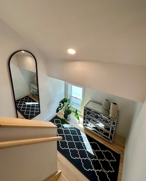 Een moderne kamer met een zwart-wit bekleed vloerkleed, een houten trap, een spiegel, een raam met uitzicht en een kleine plant.