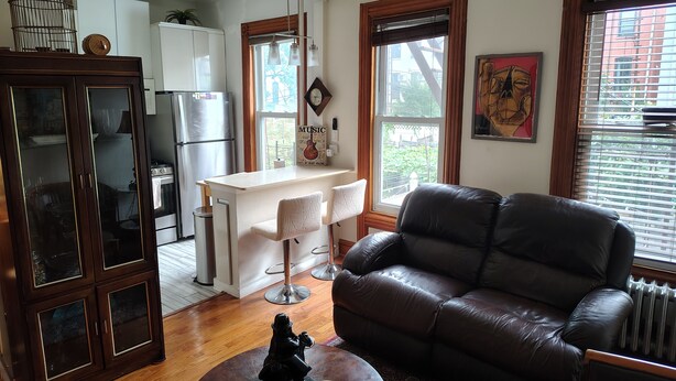 A living room with a leather sofa, a dining area with a kitchen in the background, and a large window with blinds. - Enjoyable 1-bedroom apartmen
t with AC in vibrant Brooklyn (Brooklyn)