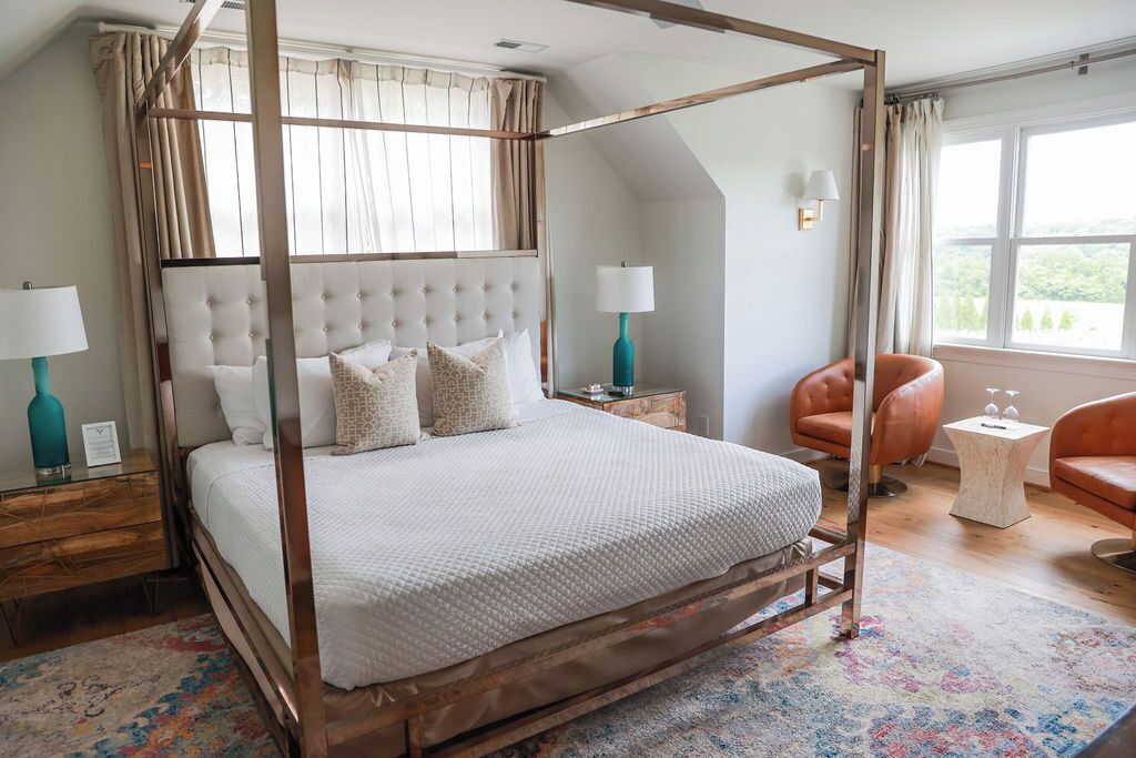 A four-poster bed with a tufted headboard, a small side table, a chair, and a view of the outdoors through a window.