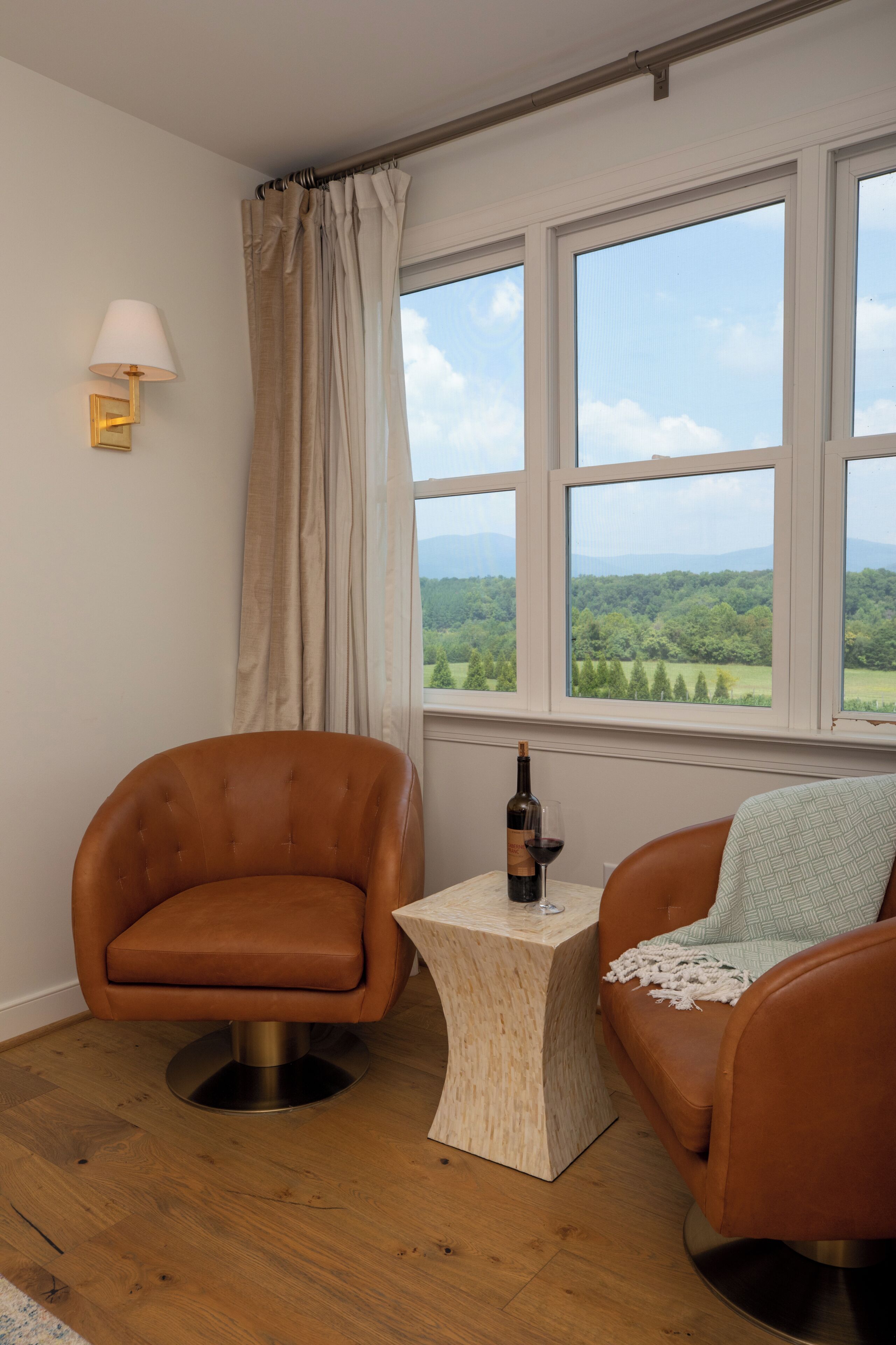 Una habitación con dos sillones de cuero, una mesita de madera con una botella y un vaso, y vistas a un paisaje verde a través de amplias ventanas.