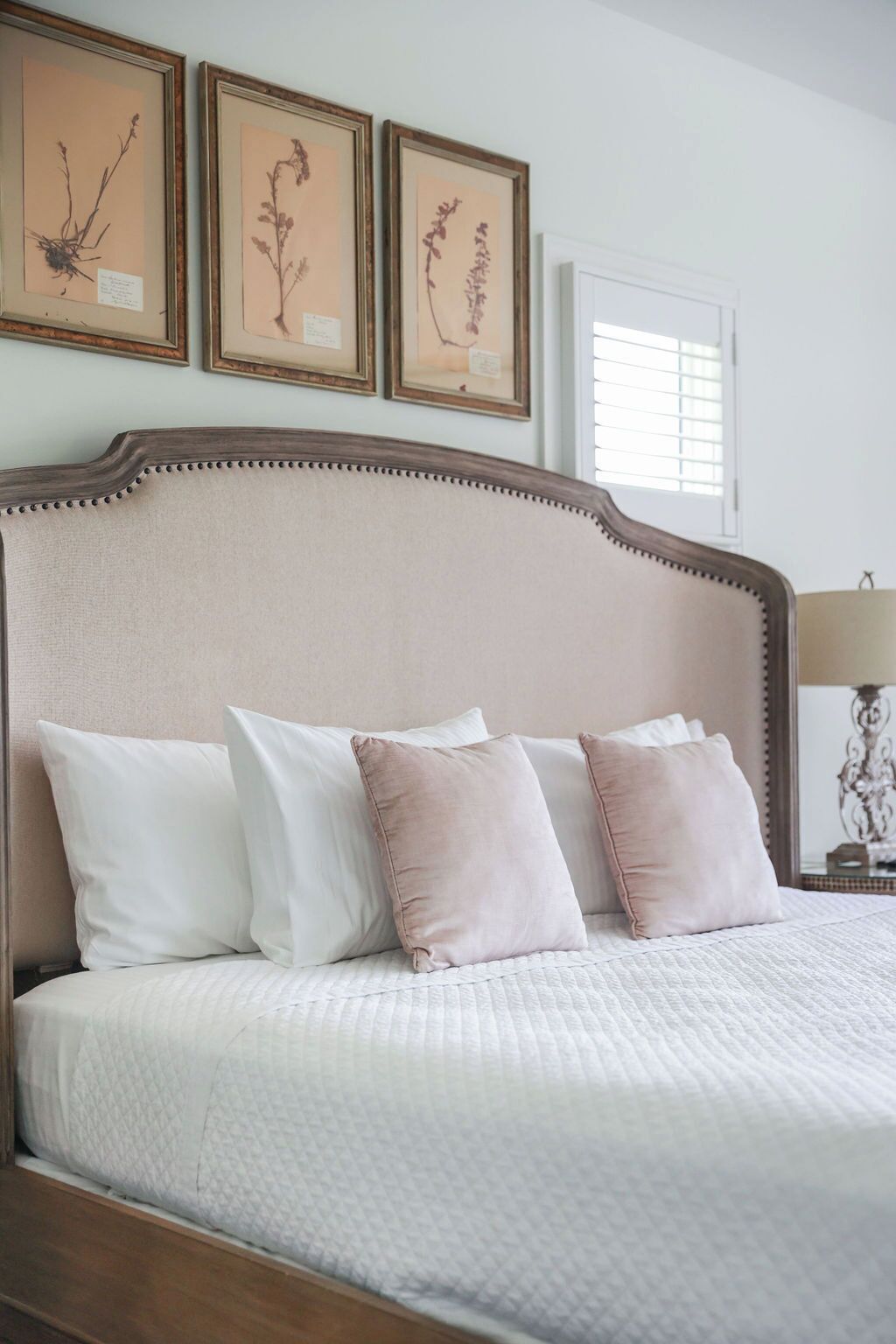 A bed with white and pink pillows, a quilted white bedspread, and a wooden headboard. There are three framed botanical prints above the bed.