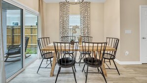 Un coin repas avec une table en bois et des chaises noires, un lustre et une vue sur le jardin à travers une porte-fenêtre coulissante.