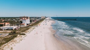 Une plage avec une jetée qui s’avance dans l’océan, des bâtiments le long de la côte et un ciel dégagé.