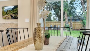 Une table à manger agrémentée d’un vase et d’une orchidée, une chaise, et une vue sur un jardin par de grandes fenêtres.