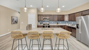Une cuisine moderne dotée d’un îlot central, de tabourets de bar, d’appareils électroménagers en acier inoxydable et de luminaires suspendus.