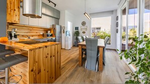 A modern kitchen with a wooden island, bar stools, a dining table with chairs, and a large window offering a view of the outdoors. - Coastal Bloom Villa Garden BBQ (Ishëm)