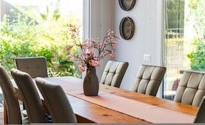 A dining table with chairs, a vase with flowers, and a window with a view of greenery. - Coastal Bloom Villa Garden BBQ (Ishëm)