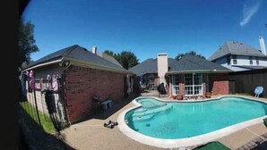 Ein Garten mit Pool, einer Pflasterterrasse und einem überdachten Bereich mit einem Stuhl.