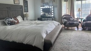 Ein Schlafzimmer mit einem großen Bett, einem Schminktisch mit Beleuchtung, einem Sessel und einem Fenster.