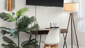 A modern office space with a desk, chair, large plant, standing lamp, and a mounted flat-screen TV.