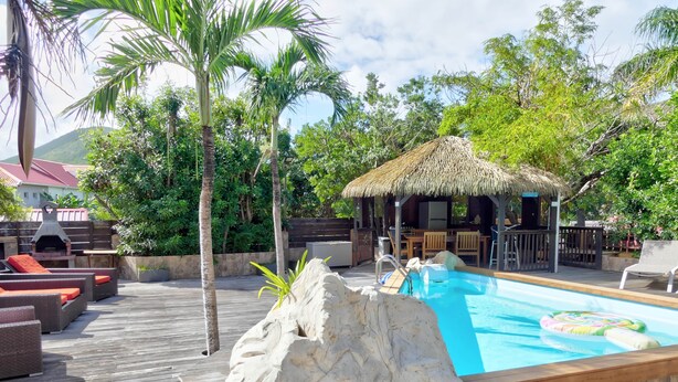 A pool area with a wooden deck, lounge chairs, a rock feature, and a thatched-roof structure. - Cozy cottage "Cocotier" w/pool in the complex (Cul de Sac)